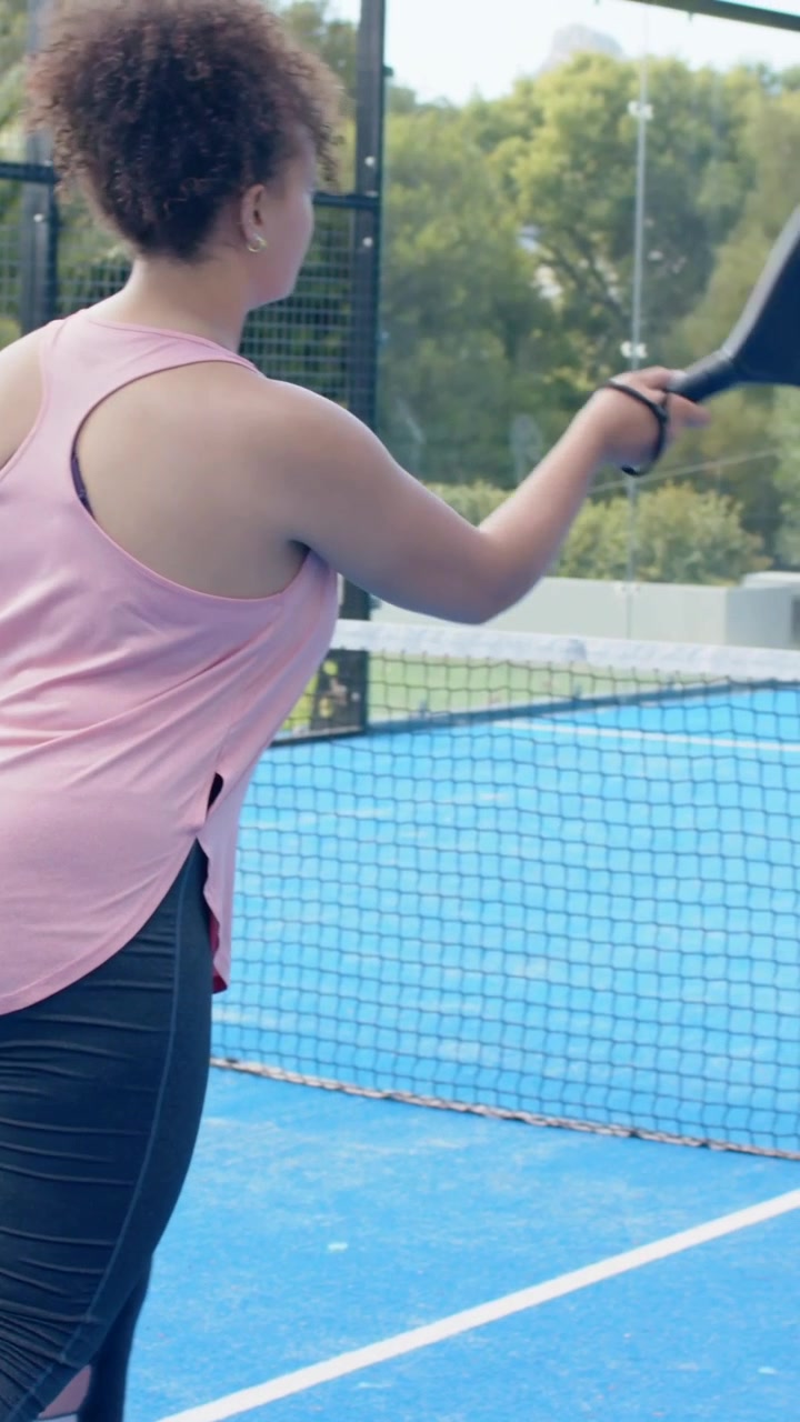Video verticale: Un amico serve la palla, una donna in rosa colpisce una palla gialla con una racchetta su un campo blu, in allenamento.