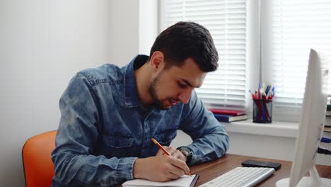 joven hombre de negocios trabajando en la computadora en una elegante oficina moderna y tomando notas usando su lápiz y cuaderno. computadora, teléfono y taza en la mesa. filmado en 4k