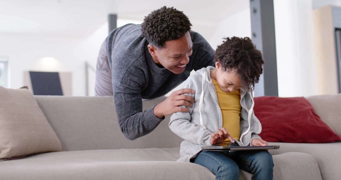 African American father and daughter leaning to see tablet on sofa, father guiding tapping for help