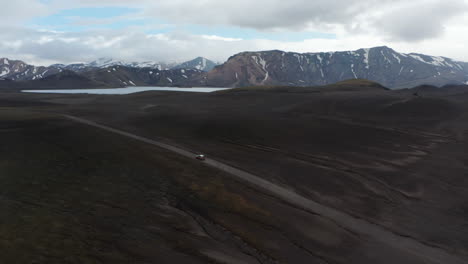 Top-view-car-driving-desert-black-volcanic-terrain-speeding-up-in-Iceland-highlands.-Aerial-drone-view-of-amazing-icelandic-countryside-with-snowy-mountains-peak-and-volcanic-black-sand-panorama
