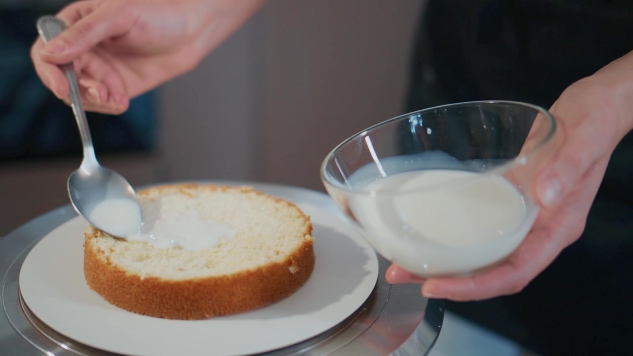 Eine Hand verteilt cremigen Zuckerguss über einen goldenen Kuchenboden. Der Kuchen steht auf einem drehbaren Ständer. Eine Schüssel enthält weiteren Zuckerguss für fortgesetztes Schichten. Eine Nahaufnahme fängt die glatte, gleichmäßige Anwendung ein.