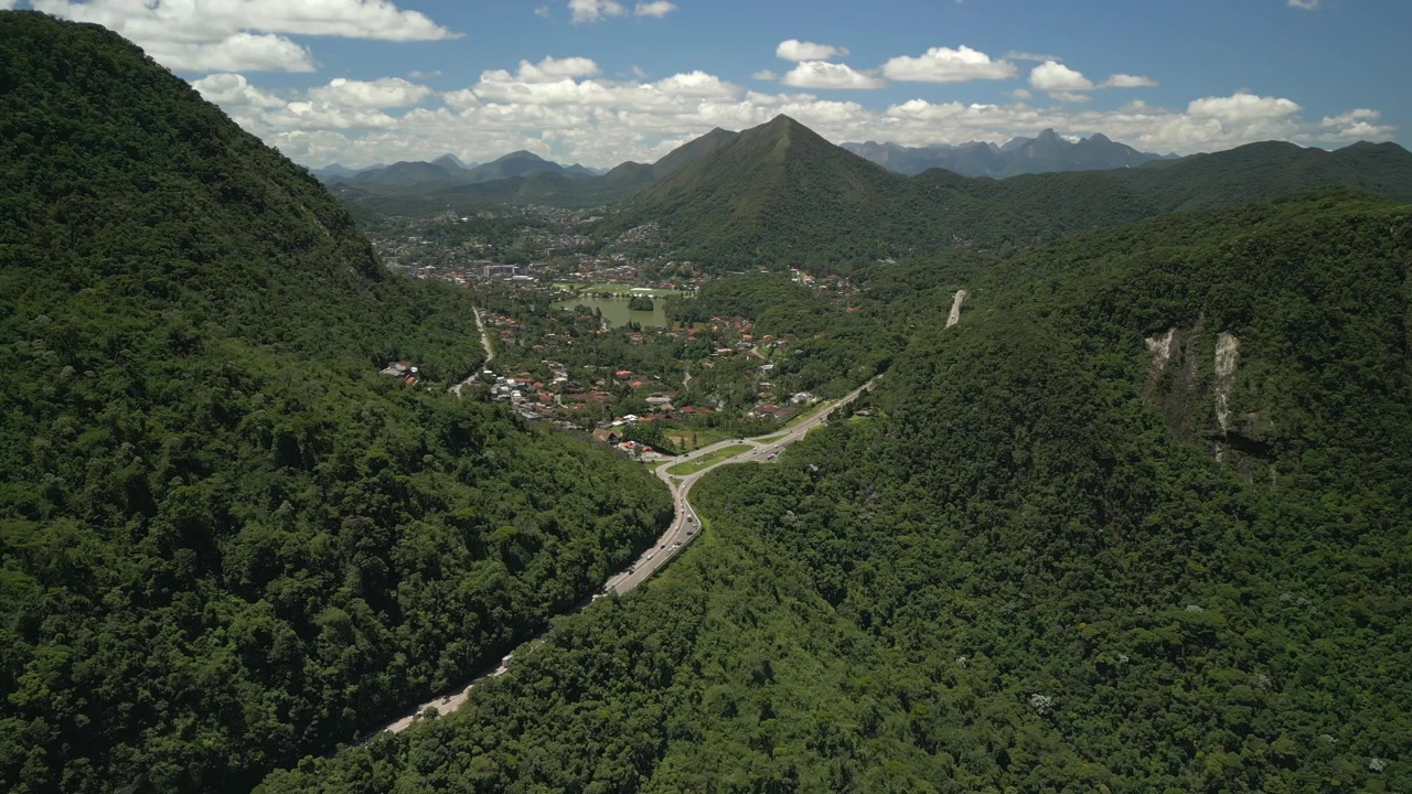 Acesso rodoviário na selva em direção a Teresópolis, com horizonte urbano emoldurado por nuvens de montanha distantes. Aproximação aérea frontal em alta altitude gravada em 4K a 25 fps