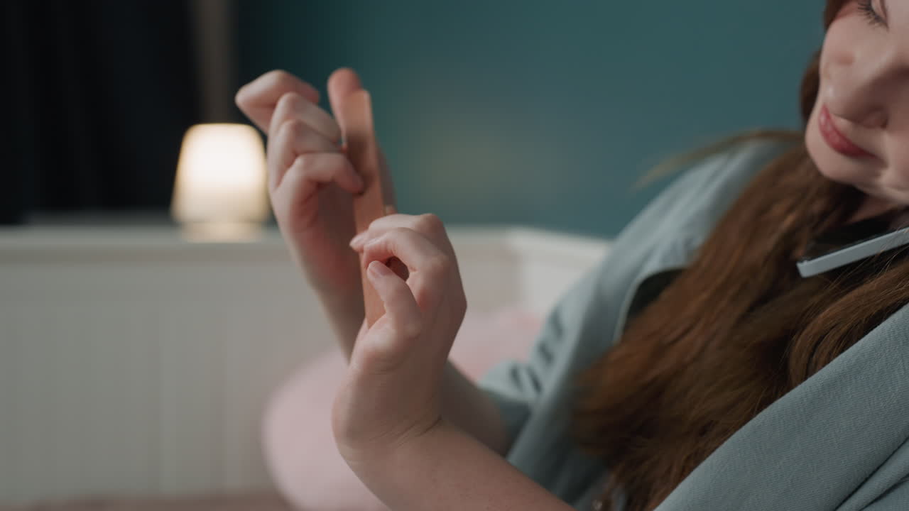 Closeup hands filing nails with emery board in soft bedroom light, intimate grooming action framed against teal wall and lamp concentrated motion, tactile texture and precision grooming evoke