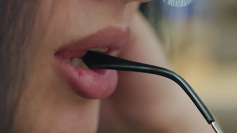 close up of young lady gently biting eyeglass arm with subtle smile on lips, showcasing soft, intimate moment with delicate facial expression and blurred warm toned background