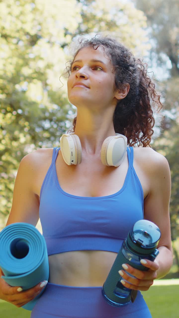 una joven deportista feliz con una alfombra y una botella de agua en las manos se dirige a entrenar al aire libre en el parque