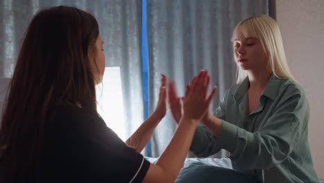 side view of young women clapping hands together while sitting on bed, playing hand game in cozy bedroom with soft lighting and relaxed mood