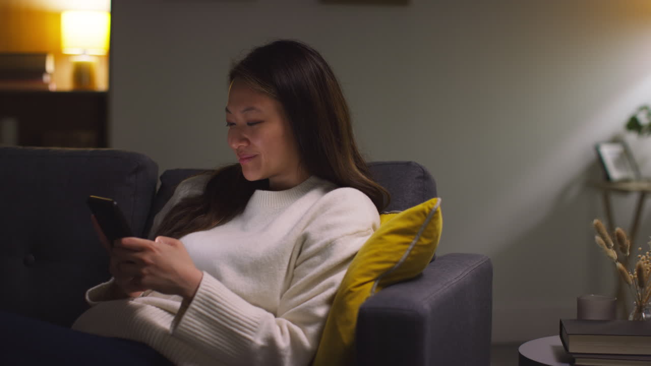 소파에서 집에서 저녁을 보내는 여성이 무선 이어폰을 착용하고 휴대 전화에서 음악이나 캐스트를 스트리밍합니다.