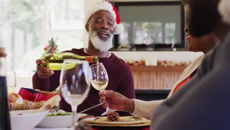 African-american-senior-man-in-santa-hat-pouring-drink-in-glass-of-senior-woman-while-sitting-on-din