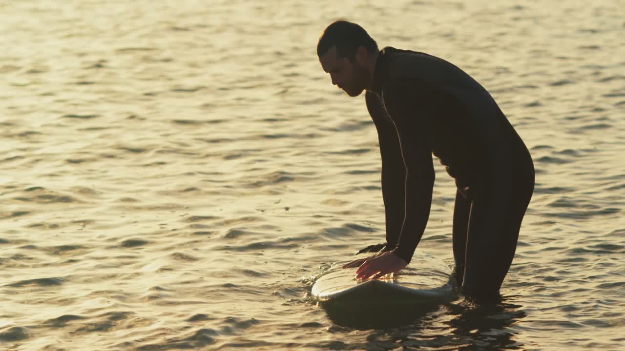 해가 지는 동안 바 ⁇ 물에서 서핑 보드를 밀고 있는 중성인 백인 남성 서퍼의 측면 뷰 4k