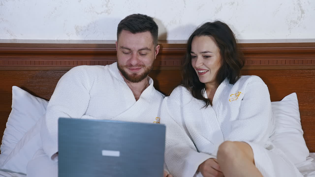 Beautiful couple in love sitting on bed and watching movie on laptop. Happy holidays of a young man and woman. Vacation and honeymoon concept.