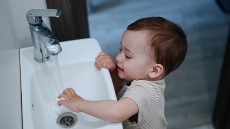happy smiling kid touches water flowing from the tap. baby boy learns to use the faucet. top view.