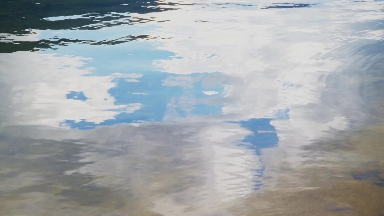 Calm water mirrors sky and clouds with gentle ripples. Surface reflects blue sky, white clouds, and distant land. Nature scene ideal for travel, environmental, or peaceful background