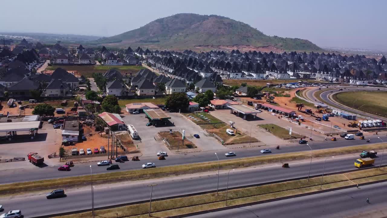 Drone voando em direção a um bairro suburbano recém-construído com casas de luxo em Abuja, Nigéria. O tráfego flui em uma rodovia próxima enquanto uma bela paisagem montanhosa pode ser vista ao fundo.