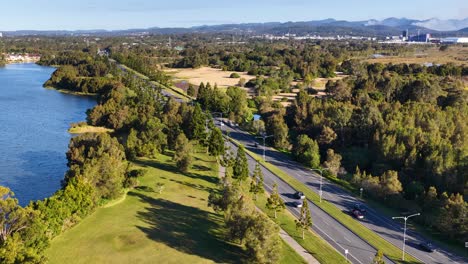 drone footage captures vehicles traveling along a tree-lined road beside a river and parkland in gold coast, australia, under clear daylight