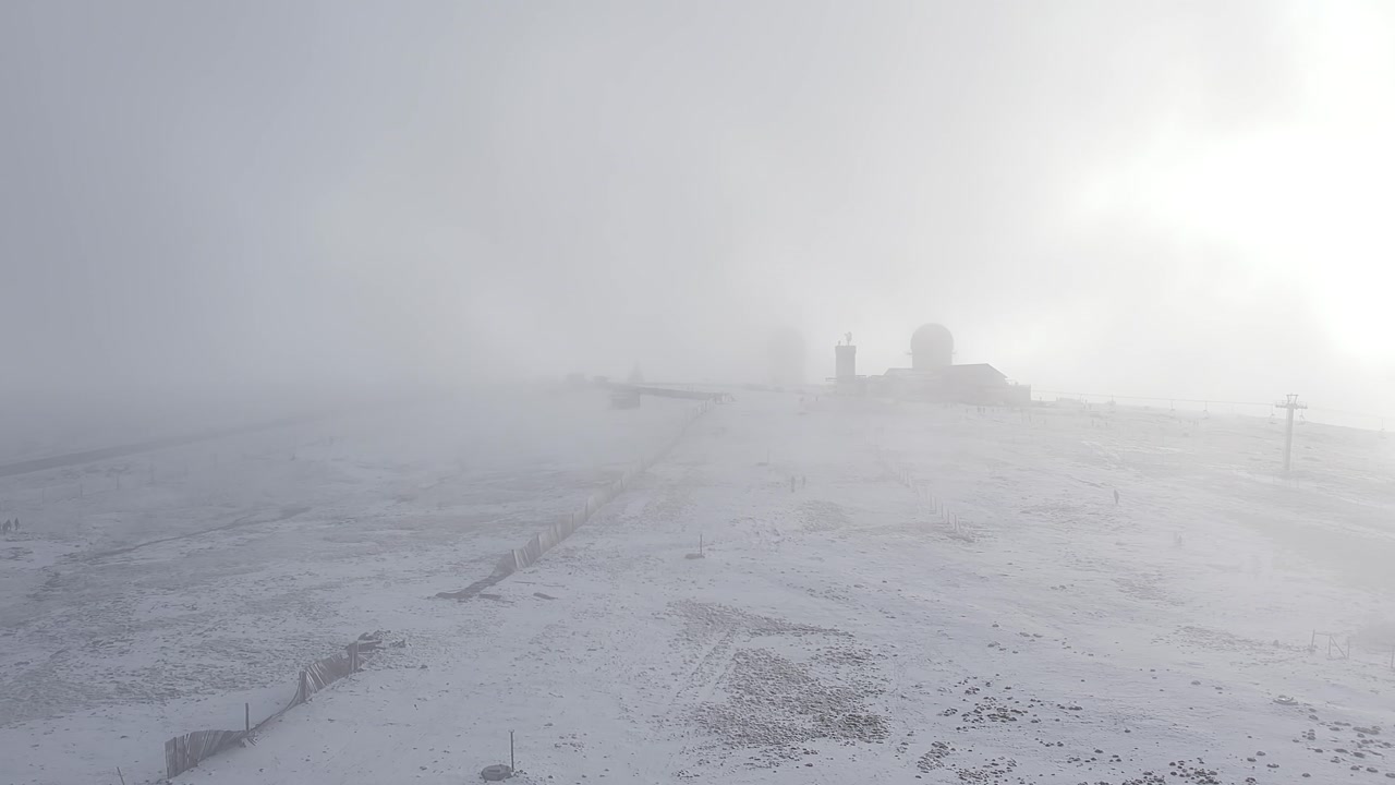 Snow-covered plateau in Serra da Estrela, Central Portugal, with dense whiteout fog and faint silhouettes of summit structures barely visible in extreme winter conditions, drone push-in