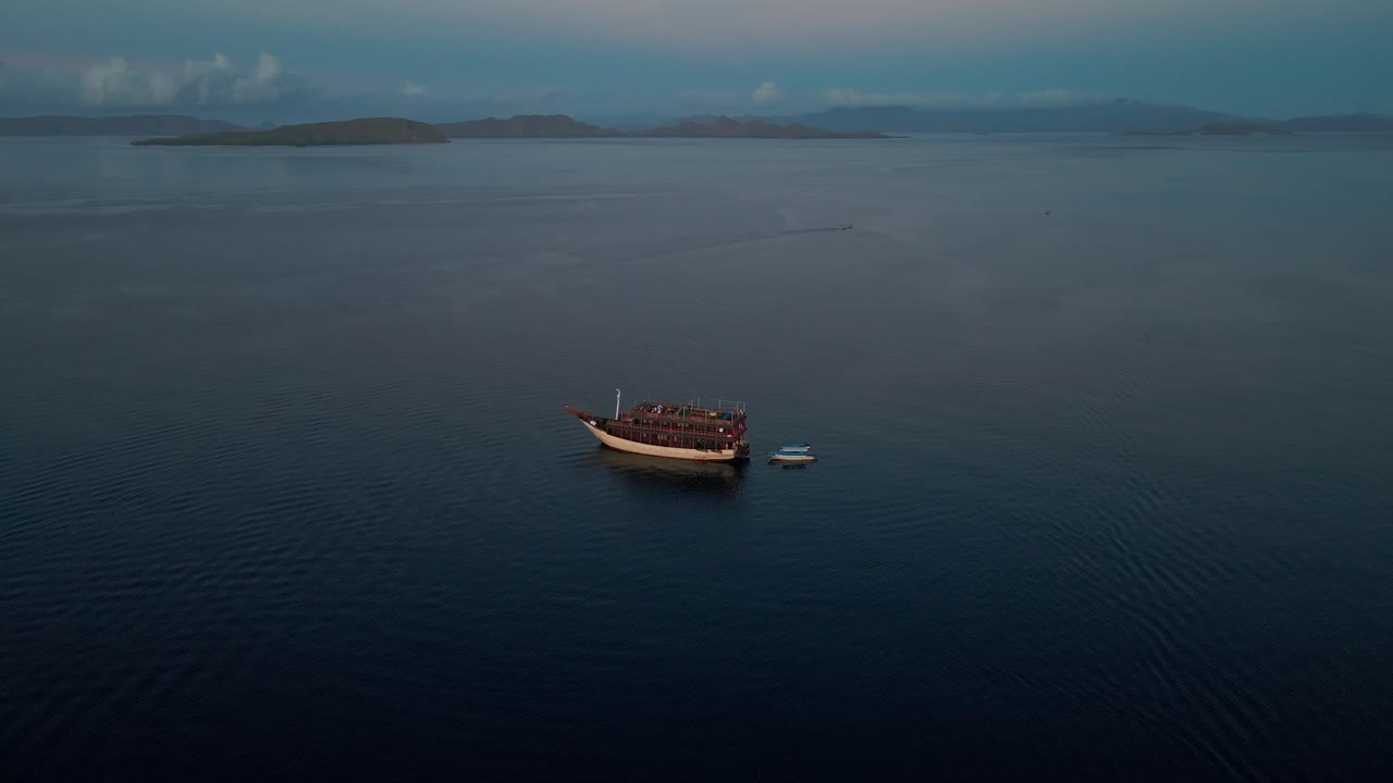 A red and white traditional boat floats on calm, deep blue Komodo waters, Indonesia reflecting the sky. In the background, a distant low-lying island enhances the tranquil scene.