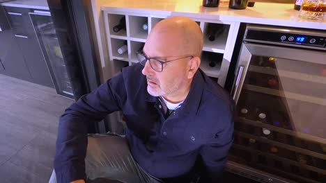 a stressed man sits on the floor in front of a bar fridge, sculling a glass of wine to ease the pain.