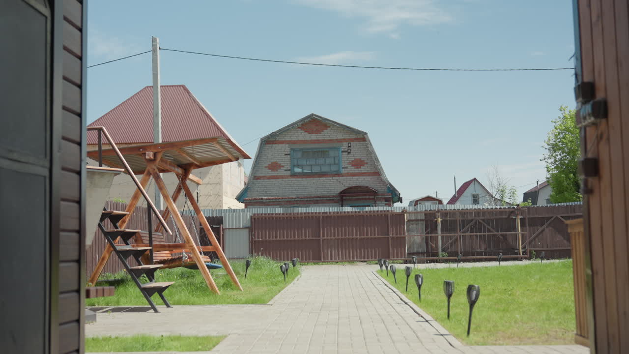 Scenic view recreational centre yard featuring wooden swing under metal roof, paved walkway lined with lights through green grass beside brown fence, suburban houses under clear blue sky