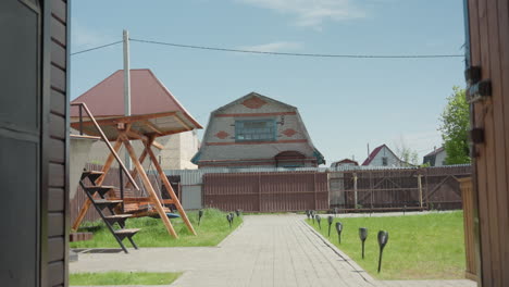 scenic view recreational centre yard featuring wooden swing under metal roof, paved walkway lined with lights through green grass beside brown fence, suburban houses under clear blue sky
