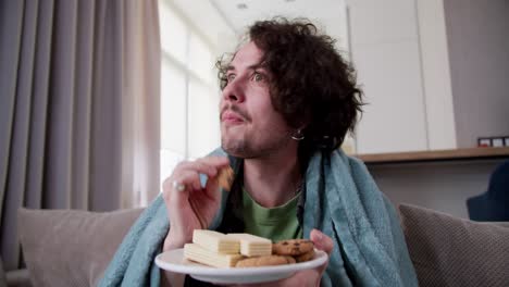 un chico moreno sorprendido y feliz con el cabello rizado está envuelto en una manta azul y come bocadillos dulces mientras ve una película interesante en casa en el sofá