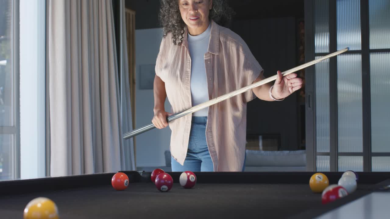 Bending woman pink-shirt lining cue, striking cue ball to sink balls on black-felt table at home
