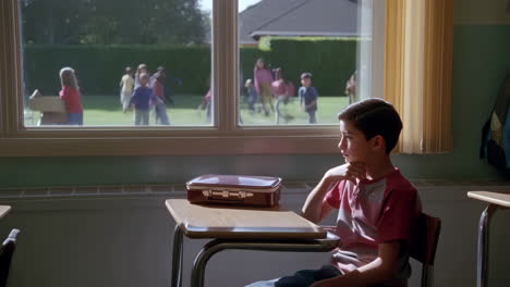 a lonely boy looks out a classroom window at children playing outside