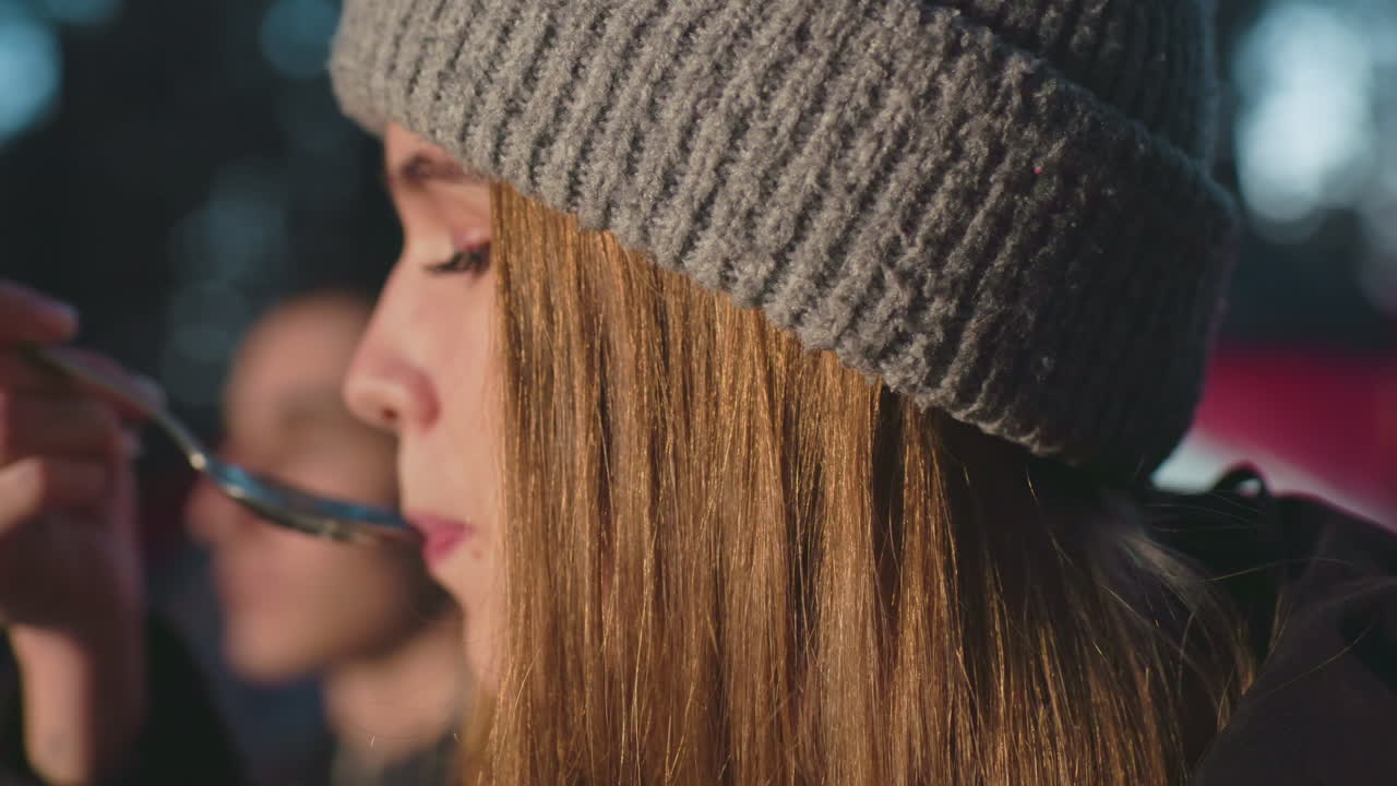 close up of young woman in knit beanie eating from spoon while seated outdoors during camping moment warm glow on face forest setting with blurred companion nearby