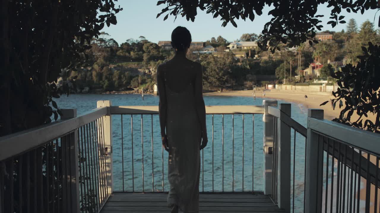 흐르는 드레스를 입은 젊은 이탈리아 여성은 물가 산책로에서 일몰을 지켜보고 있습니다.