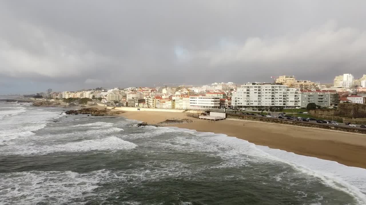 Vista aérea con dron de las olas del océano rompiendo en una playa de arena con edificios alineados a lo largo de la costa en Póvoa de Varzim, Portugal, mostrando el paisaje urbano costero bajo un cielo nublado dramático
