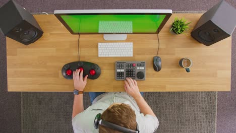 overhead view of frustrated male video editor wearing wireless headphones and working at computer with green screen in creative office - shot in slow motion