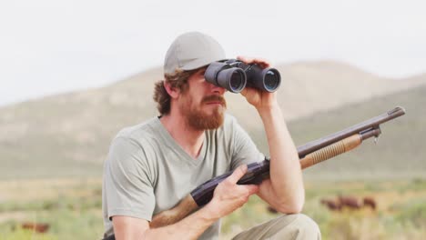 sobreviviente caucásico barbudo que sostiene un rifle de caza buscando con binoculares en el desierto