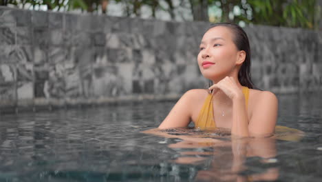 dreaming asian woman leaning chin on hand at the edge of swimming pool and looking aside smiling, thailand resort, face closeup slow-motion