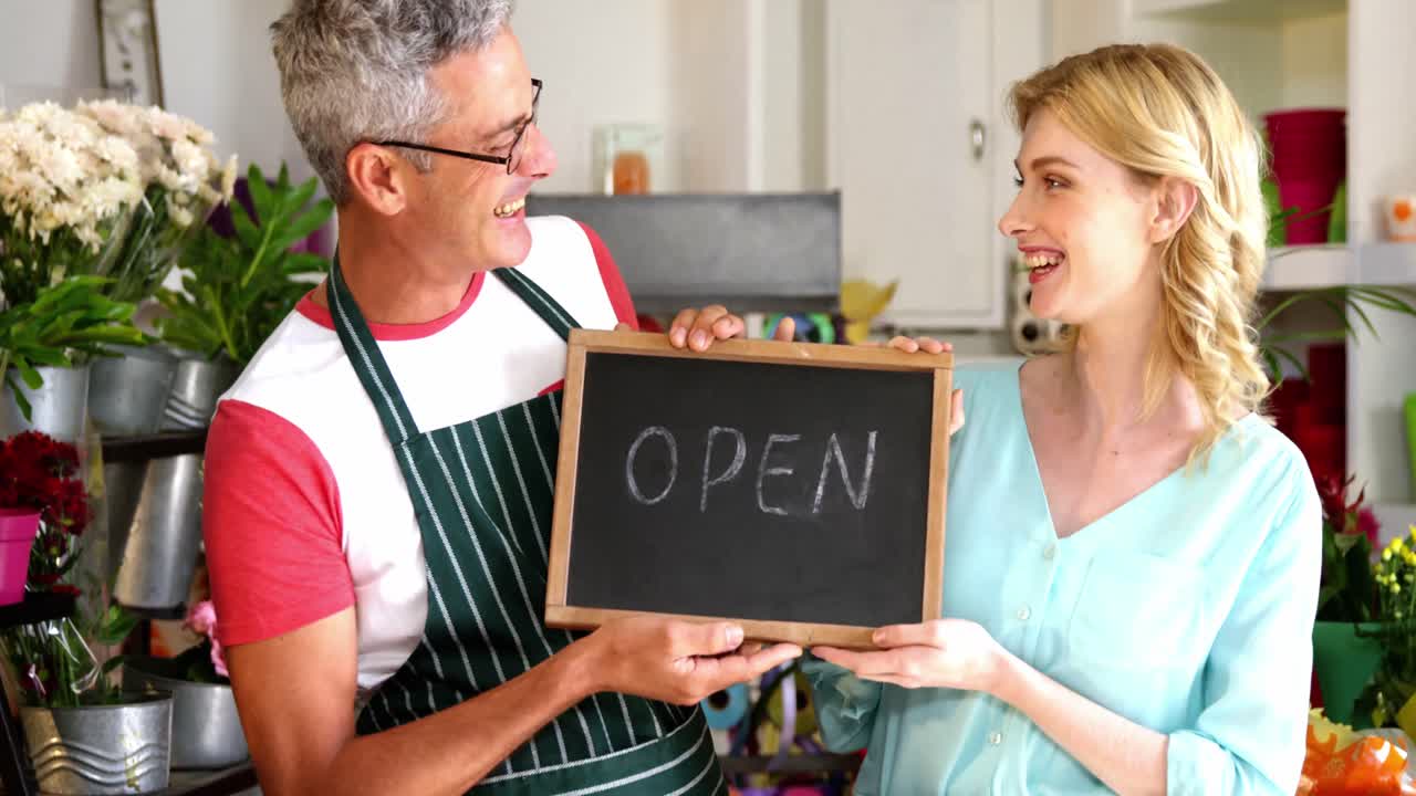 Smiling florists holding open sign on slate in flower shop