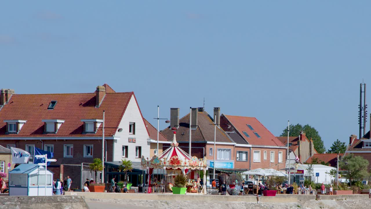 Colorful carousel rotates beside beach town buildings under bright daylight, wide static camera shot