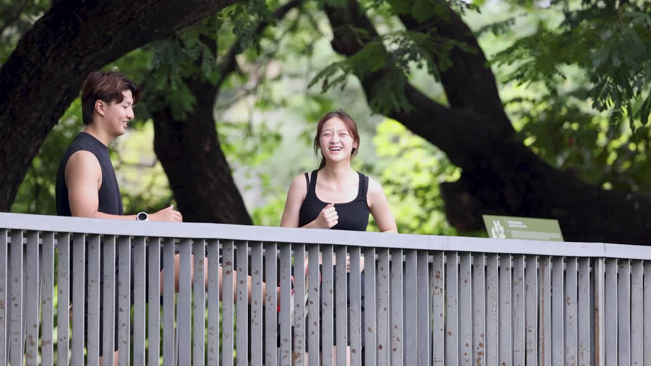 คู่รักชาวไทยที่แข็งแรงแข็งแรงวิ่งเหยาะๆ ข้ามสะพาน หัวเราะด้วยกันภายใต้ต้นไม้เขียวชอุ่มในสวนสาธารณะ