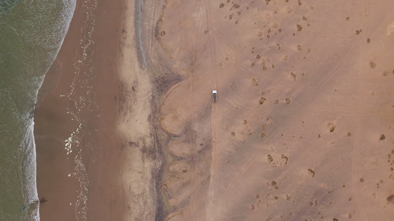 Top down drone footage following an off-road vehicle driving along the sandy beach near Swakopmund, Namibia, under overcast skies