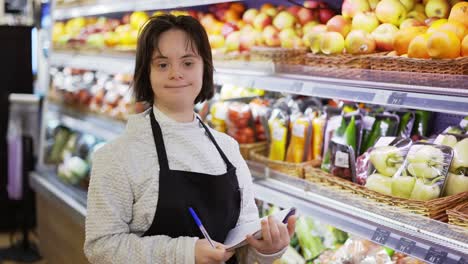 Girl-with-Down-syndrome-inspecting-shelves-with-fresh-fruits-in-a-grocery-store-using-notebook