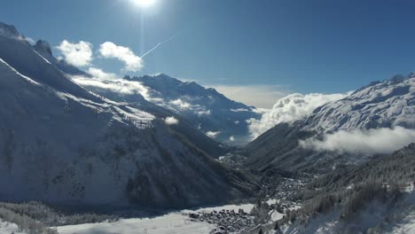 Drone-Footage-Over-Snowy-Valley-with-Distant-Mountains,-Sunlit-Sky,-and-Tree-lined-Slopes