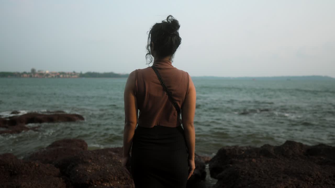 A woman stands on rocky shore gazing at the ocean during sunset