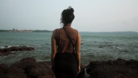 a woman stands on rocky shore gazing at the ocean during sunset