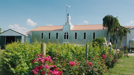 a large agricultural hangar with roses in the foreground and a small wind generator in the back grap