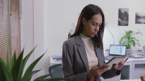 mujer de negocios biracial sonriente sentada en un escritorio usando una tableta en una oficina moderna vacía