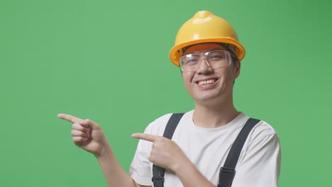 close up of asian man worker wearing goggles and safety helmet smiling and pointing to side while standing in the green screen background studio