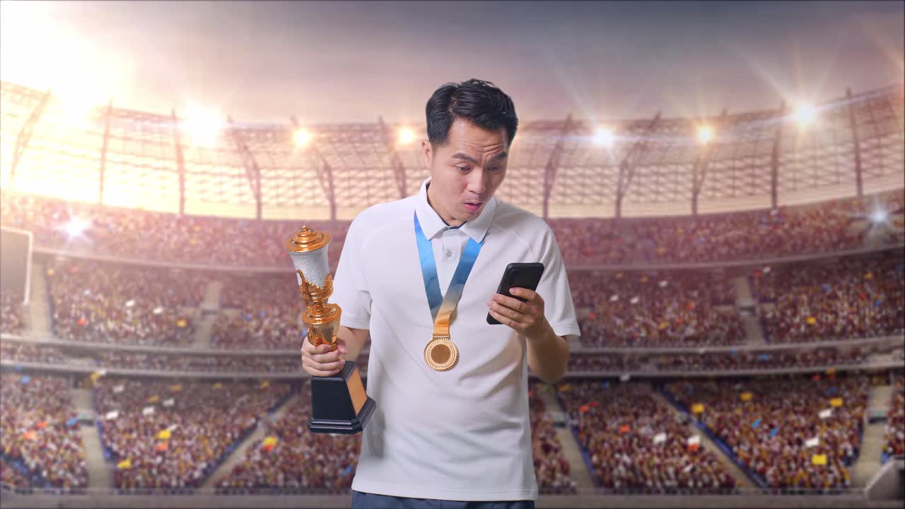 금메달과 트로피를 가진 아시아인 남자가 스마트폰을 보고 스타디움에서 와우라고 말합니다.