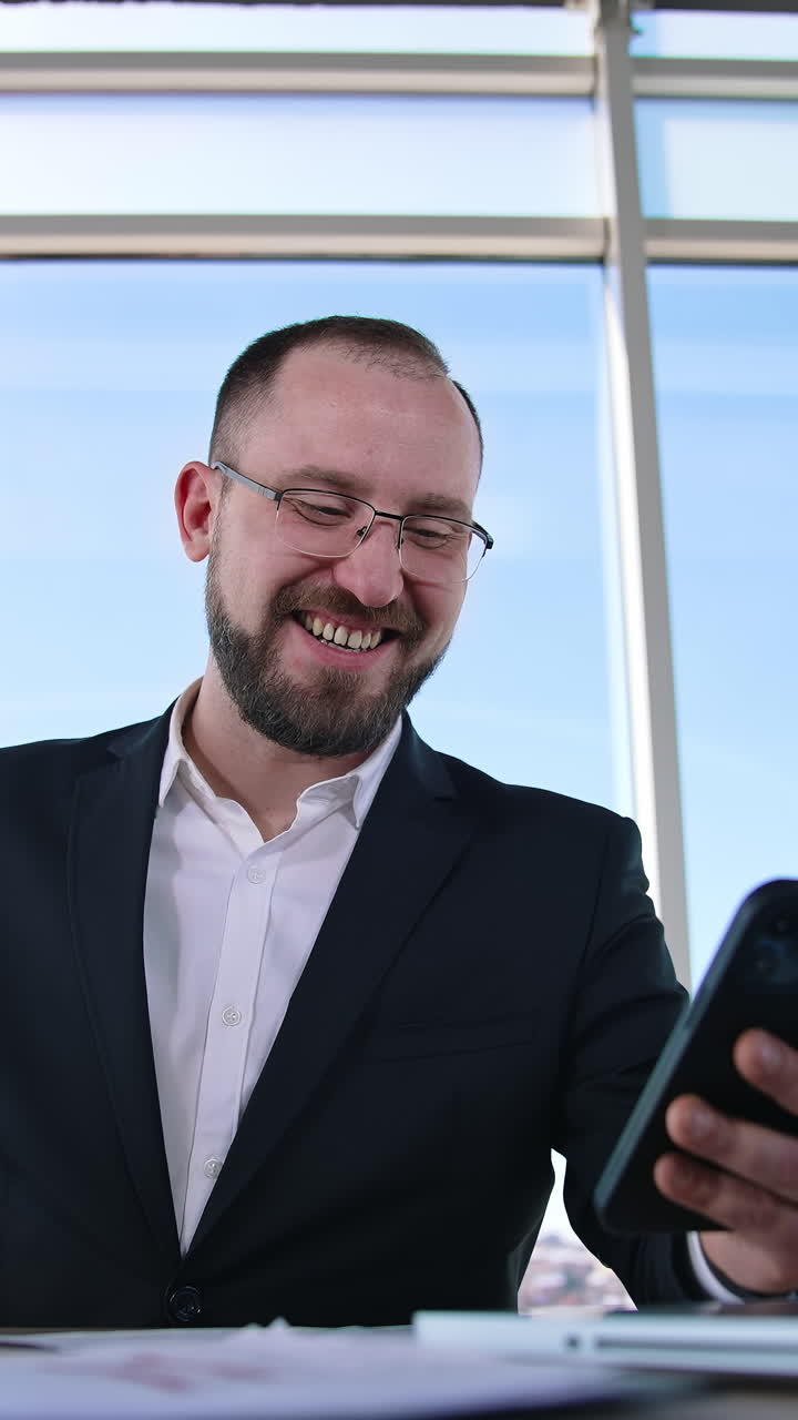 Smiling businessman having a video call from his phone. Office worker in glasses wearing suit sitting at table with a laptop using mobile phone. Vertical video