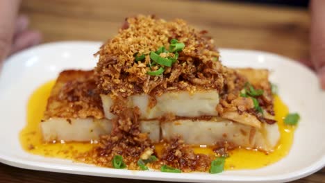 a plate of fried turnip cake garnished with crispy shallots and green onions is presented on a wooden table under warm, even lighting, with gentle camera movement