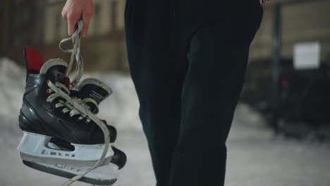 front view of person in winter outfit walking on icy surface holding black ice skating boots in right hand with left hand inside pocket, snowy surroundings and soft city background