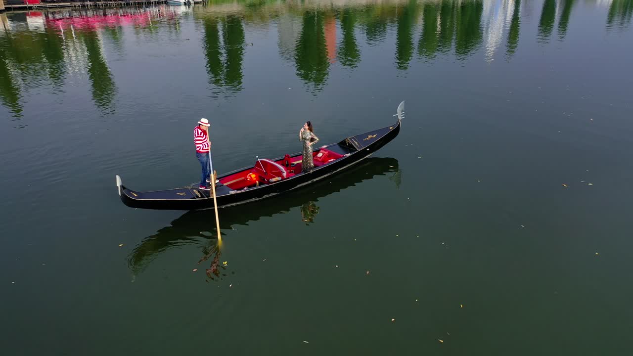 Gondolier riding a boat with a woman. Beautiful lady travelling in gondola along the river at sunset. Aerial view. Camera motion around.