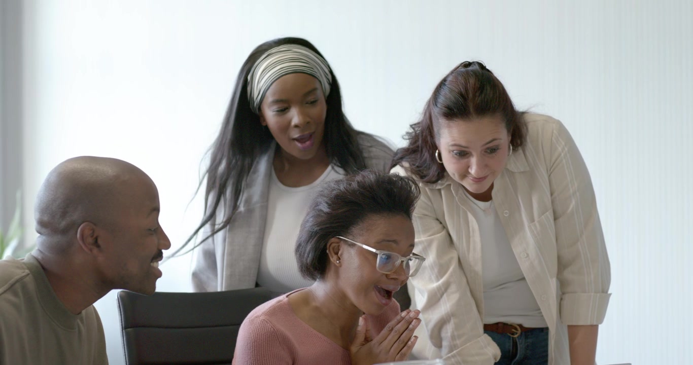 Equipe diversa reagindo ao laptop, mulher afro-americana apontando, colegas se unindo para celebrar na mesa.