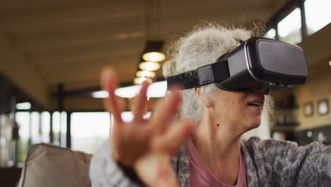 Senior-mixed-race-woman-sitting-on-sofa-wearing-vr-headset-touching-virtual-screen